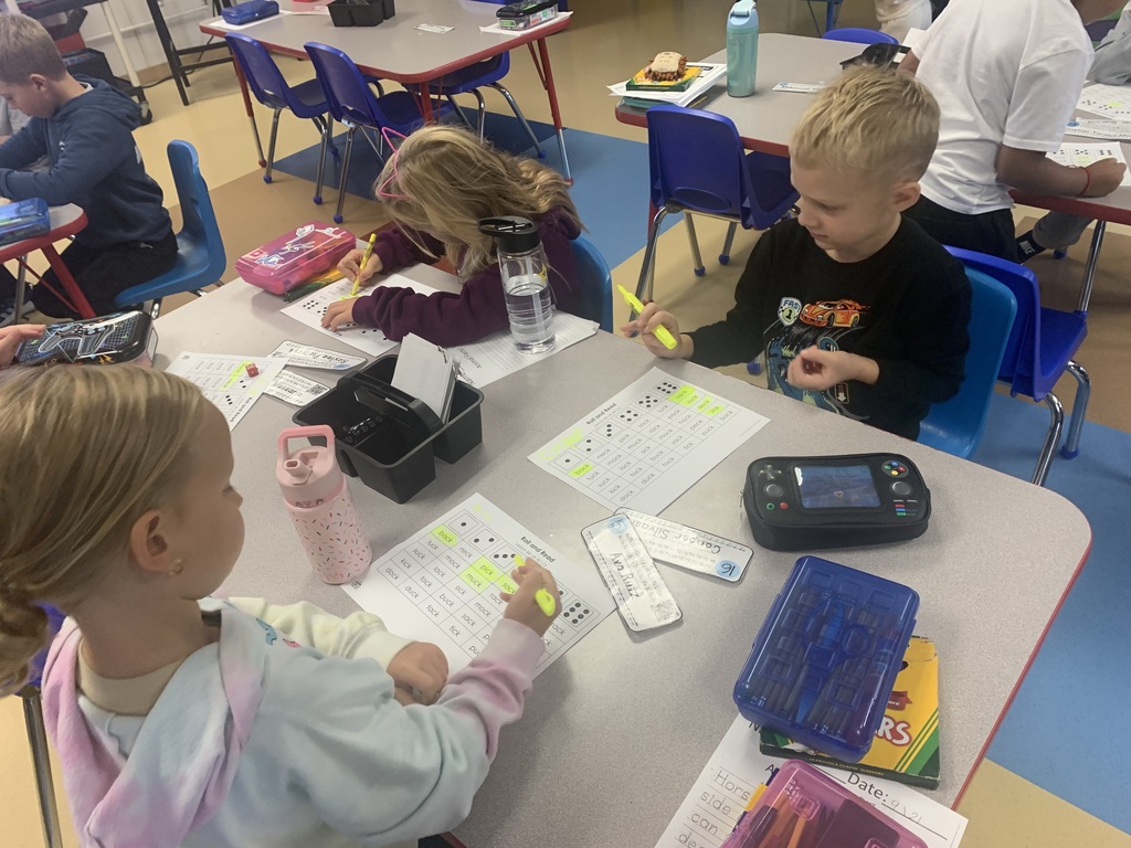 Young elementary students sitting at a classroom table working on reading worksheets with highlighters and dice.