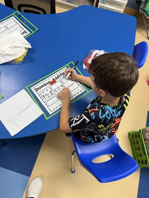 A young student sitting at a blue classroom table writing answers on a domino addition math activity sheet.