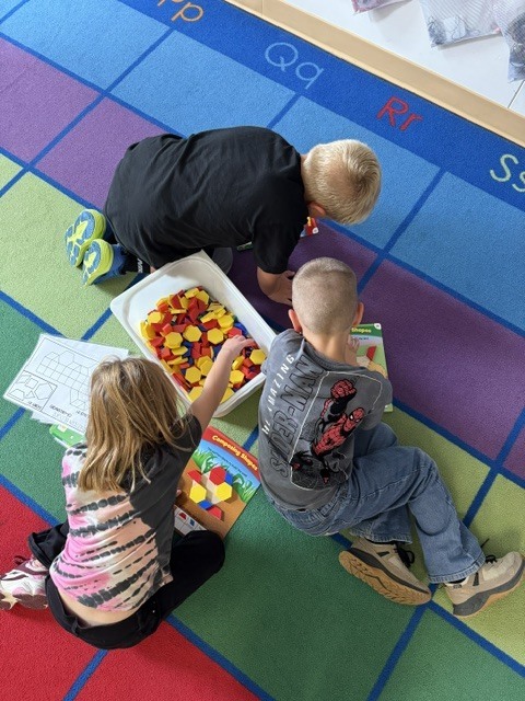 Elementary students lying on an alphabet rug working together with colorful pattern block math manipulatives.