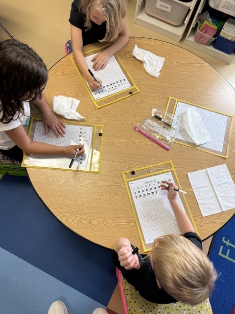 Four young students sitting around a round wooden table filling out math worksheets in reusable dry-erase sleeves