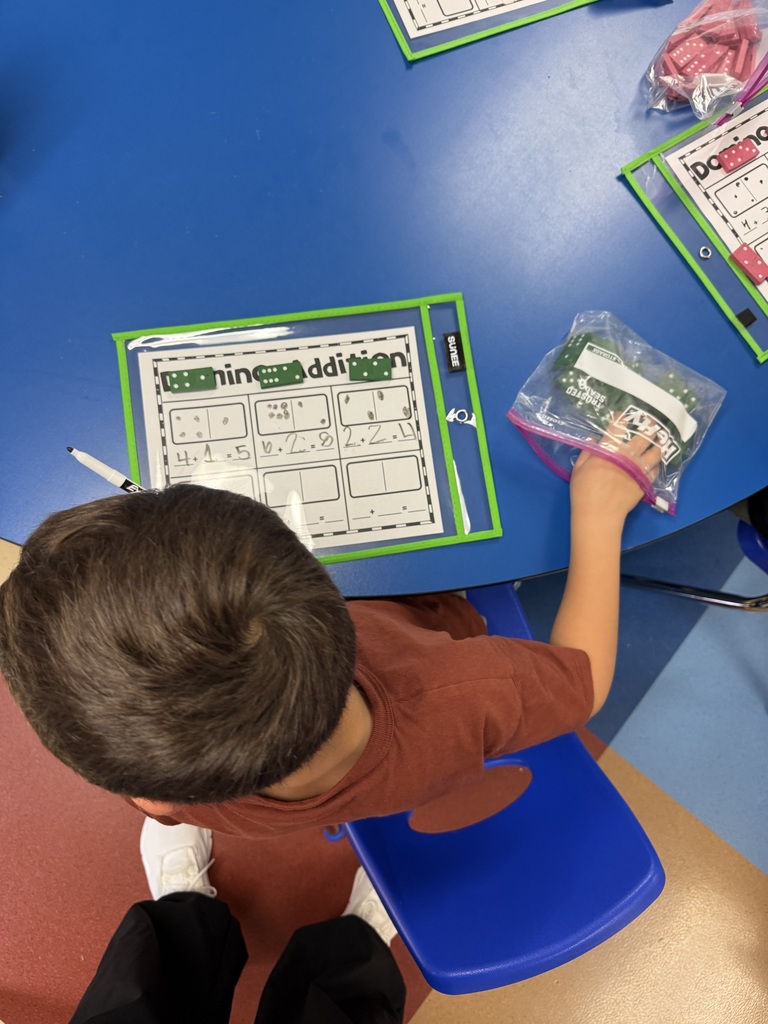 An overhead view of a student working on a domino addition worksheet at a blue table with a bag of green dice.