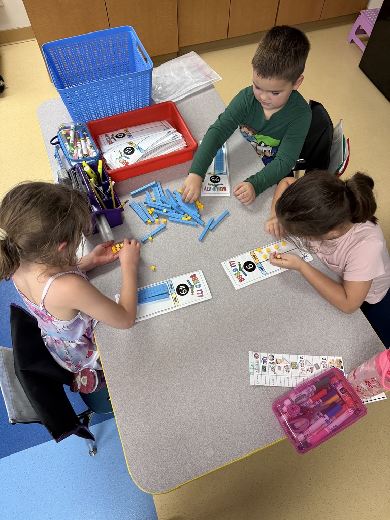 Three elementary students sitting at a classroom table using base-ten place value blocks on "Build It!" math mats.