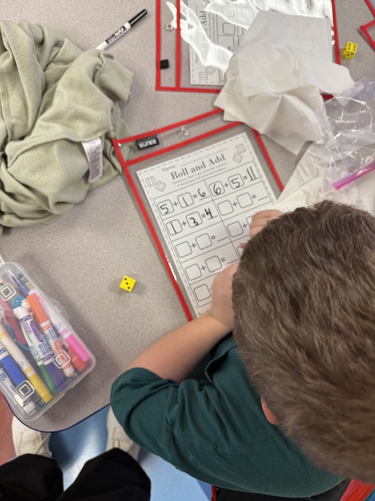 An overhead view of a student filling in addition equations on a "Roll and Add" worksheet using a die.