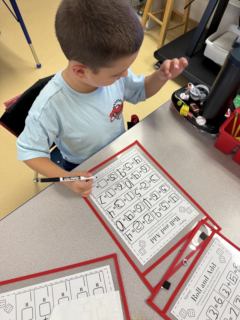 A young boy sitting at a desk completing a dry-erase math addition worksheet with a dry-erase marker.