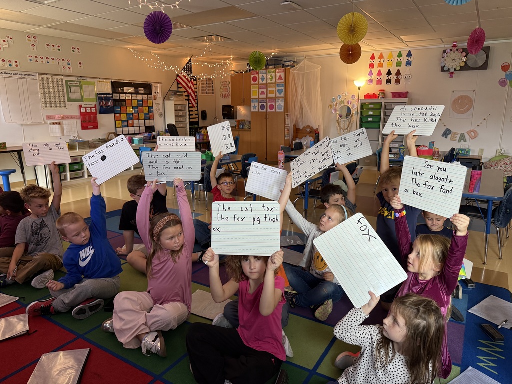 Classroom students sitting on a colorful carpet holding up lined dry-erase boards to show their writing work.