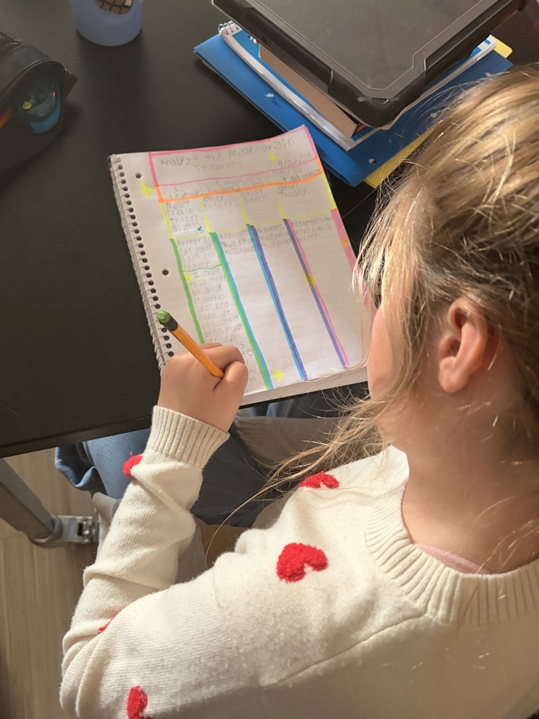 Over-the-shoulder view of a student wearing a heart sweater writing in a color-coded notebook with a pencil