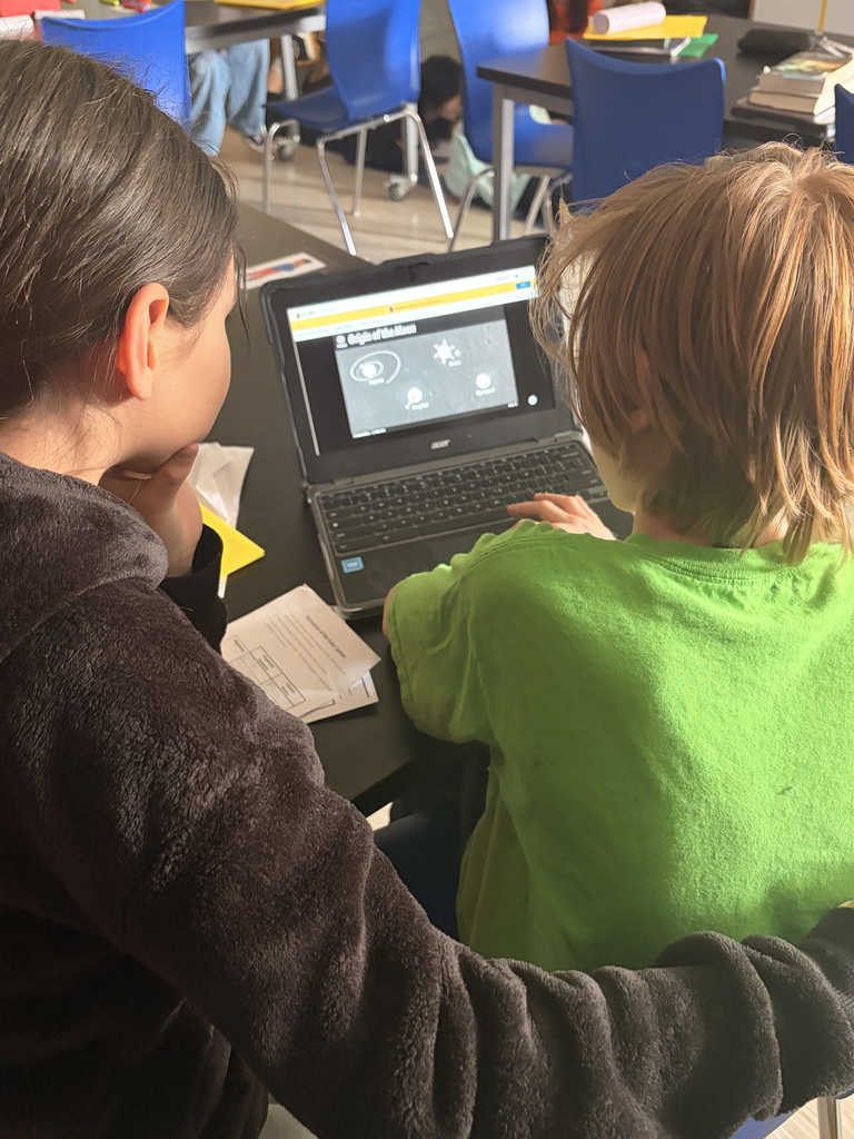 Two middle school students sit together at a classroom table looking at an educational video on a laptop screen