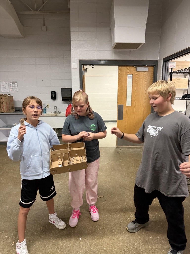 Three middle school students collaborate and examine a multi-room cardboard building model together.