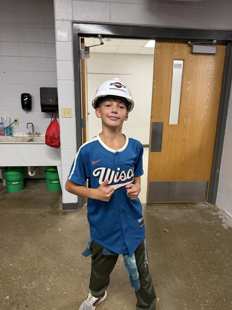 A young student wearing a white construction hard hat and blue sports jersey gives two thumbs up.