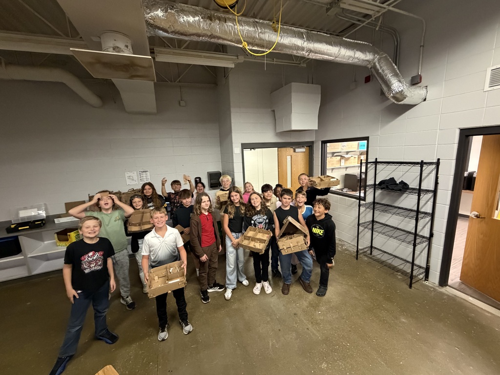 A large group of middle school students pose together, proudly displaying handmade cardboard structure projects