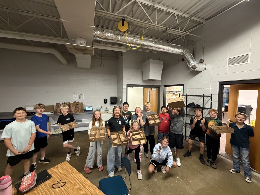 A group of students gather in a classroom holding up cardboard house models and engineering projects.