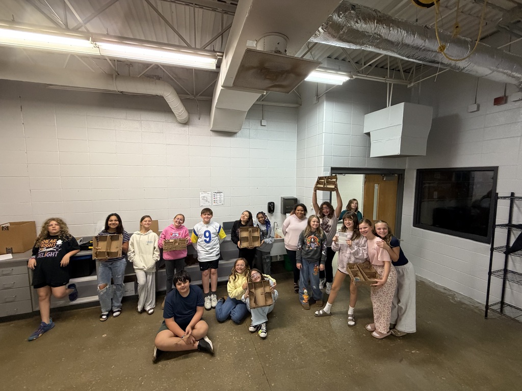 A group of students gather in a classroom holding up cardboard house models and engineering projects.