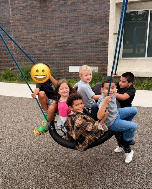 A group of elementary students sitting together on a large round web swing outdoors.