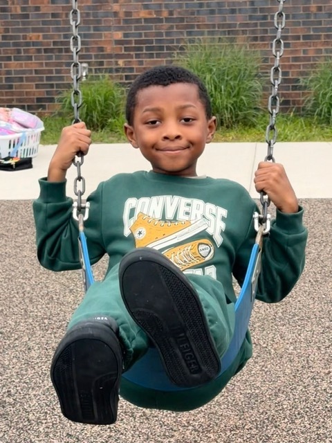 A young student smiling on a playground swing while wearing a green sweatshirt.