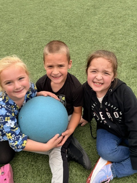 Three students laying together on artificial turf with a blue playground ball.