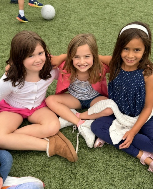 Three young students sitting side-by-side with arms around each other on grass outdoors.