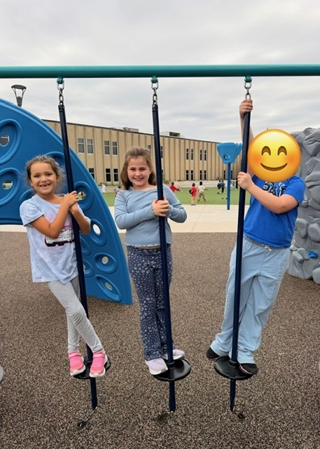 Three students balancing on vertical playground poles outdoors.