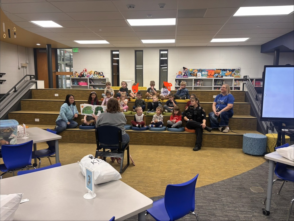 Ms. Krolow holds up a picture book in front of young students sitting on tiered steps in a school media center.