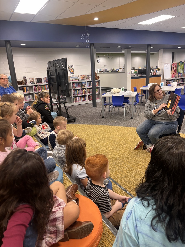 Young students sit on colorful cushions listening intently as MS. Krolow reads a book aloud.
