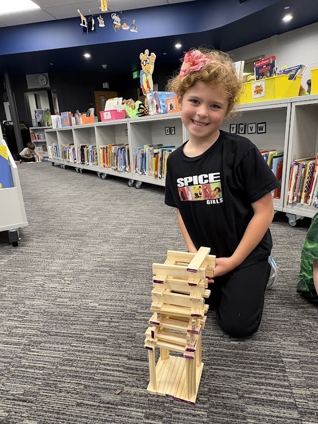 A young student wearing a Spice Girls shirt smiles next to a wooden plank tower in the school library.