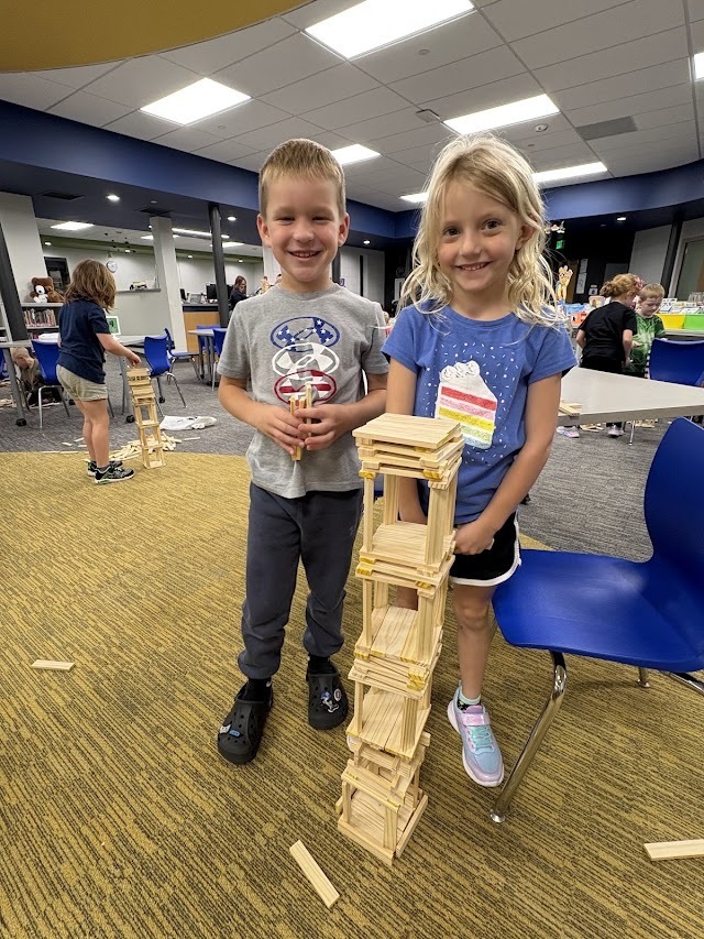 Two students smile next to a multi-level wooden block tower built on the classroom floor.