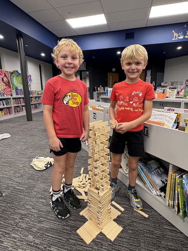Two smiling boys stand on opposite sides of a tall wooden plank tower built in the school library.