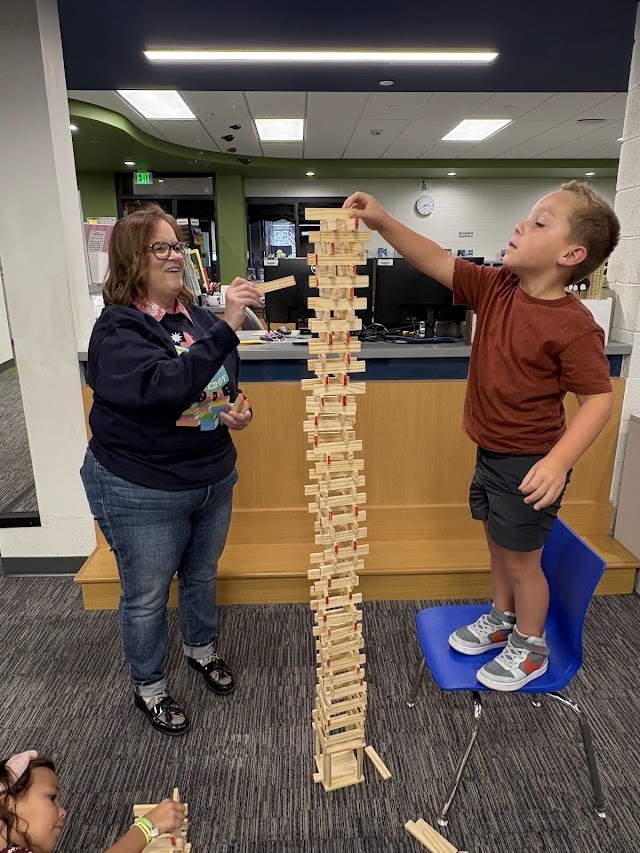 student standing on a blue chair carefully places a plank on top of a tall wooden block tower assisted by a teacher.