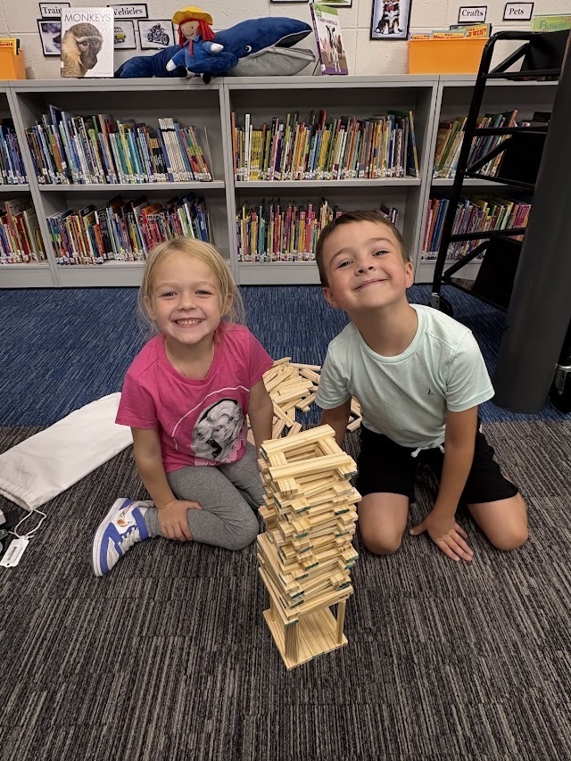 Two smiling elementary students kneel on the floor next to a tall wooden block tower in a school library.