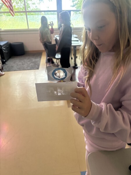 A student demonstrates how light focuses through a paper eye model onto a paper retina screen in a classroom.