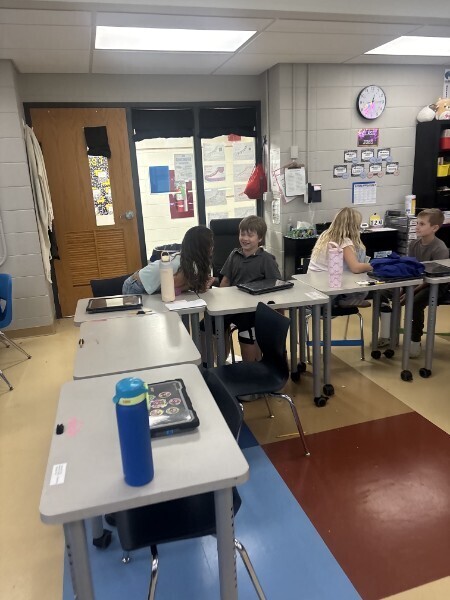 Students sit at desks working on laptops and chatting together in a classroom setting.