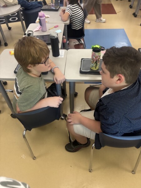 Two students sit facing each other across desks during an interactive classroom discussion activity.