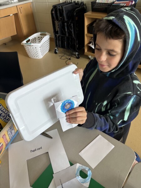 A student holding a paper eye model and lens card demonstrates an optical science experiment at their desk