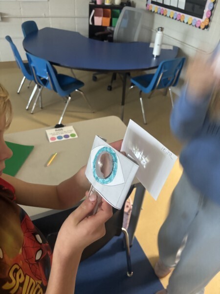 A student holds a paper eye model and a card labeled "retina" during a science experiment in a classroom