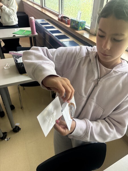 A student holds up a paper eye lens model and white paper card to observe projected light during a science lesson