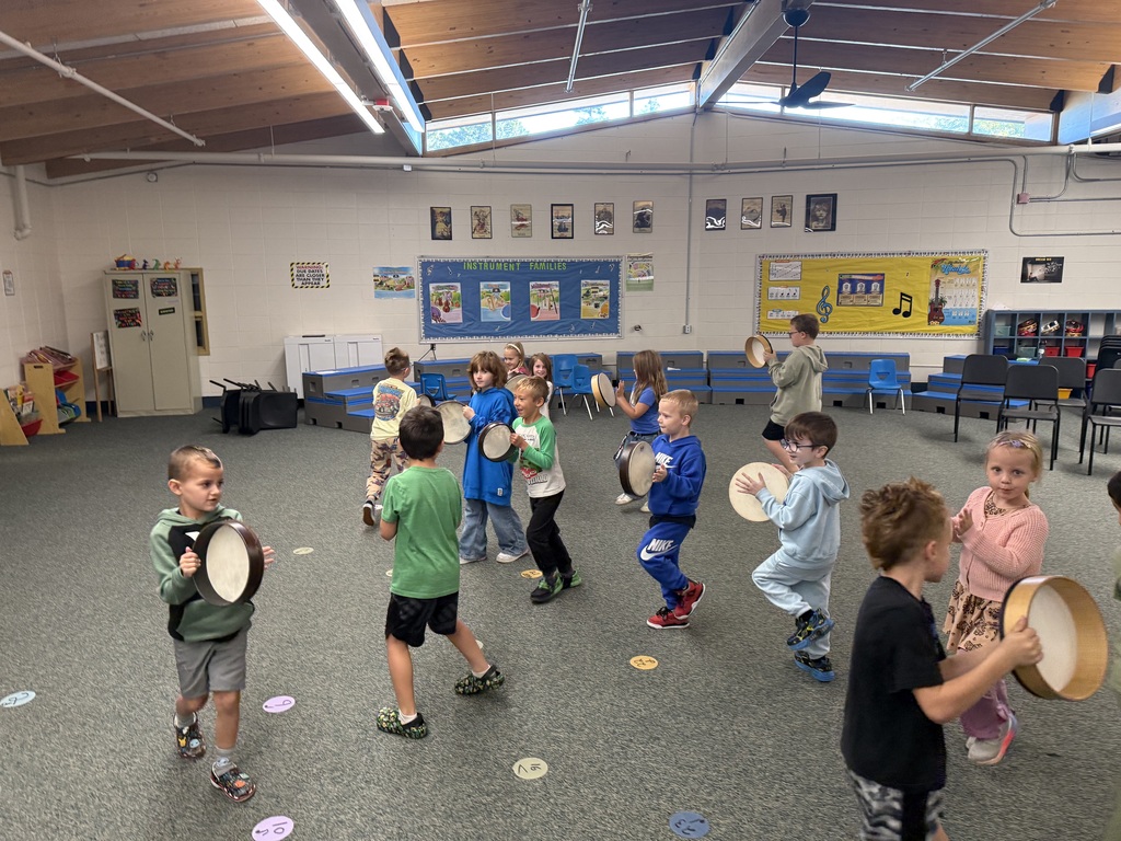 Elementary students dancing and playing hand drums in a circle around their music classroom.