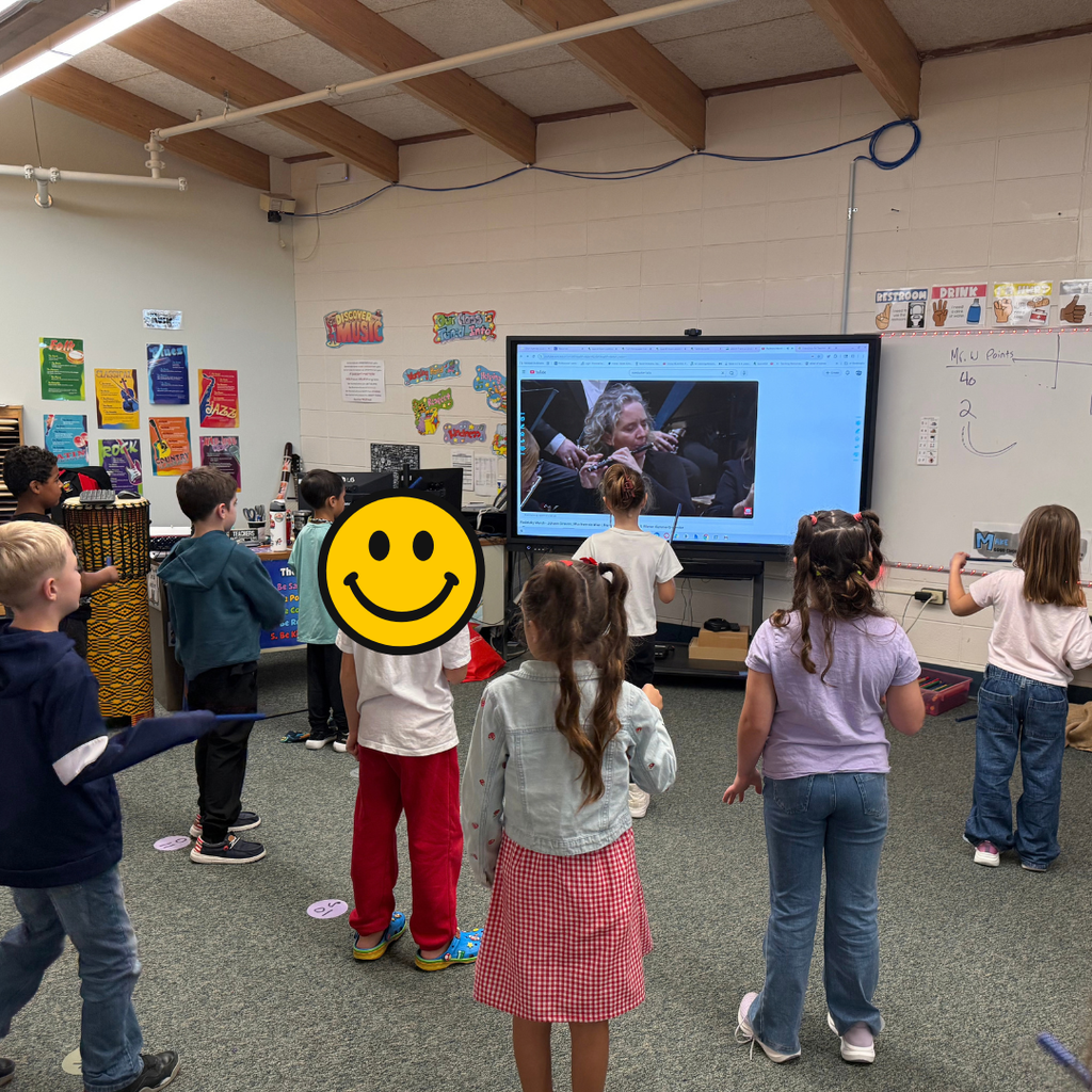 Children in a music room facing an interactive monitor while practicing rhythm movements with drumsticks.