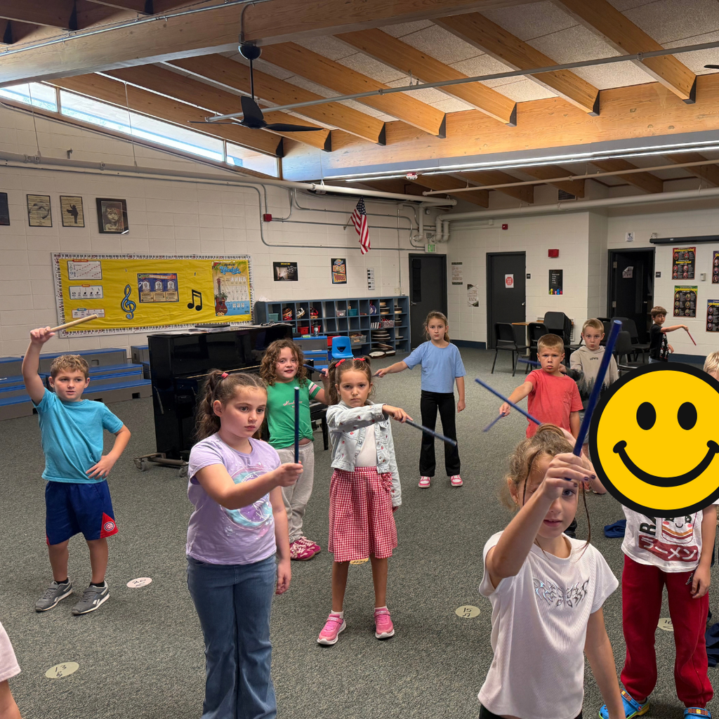 Elementary music students holding up blue drumsticks while posing together in their classroom.
