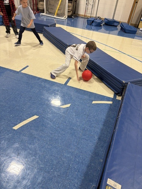 A student in a baseball jersey stoops to pick up a red ball in the school gym.