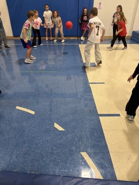 Students gathered in a school gymnasium playing a game with a red ball during physical education class.