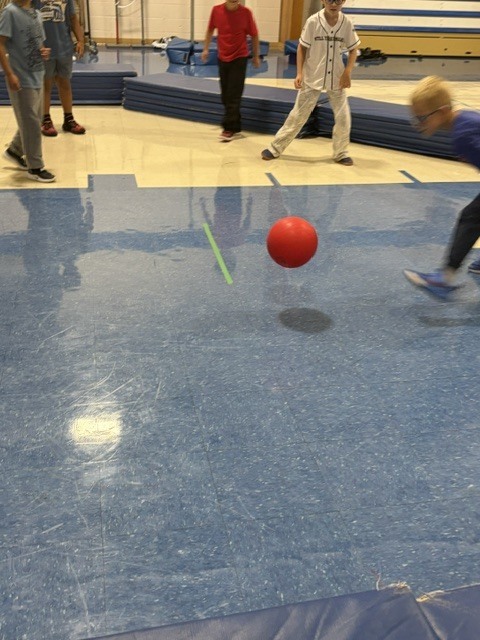 A red ball rolls across the gymnasium floor while students play during gym class.