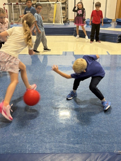 Students kicking and dodging a red ball while playing a game in the school gymnasium.