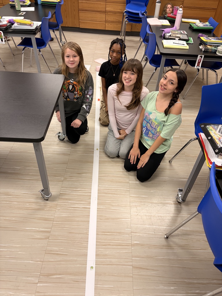 Four students kneeling around a long paper tape measure on the classroom floor during a group activity.