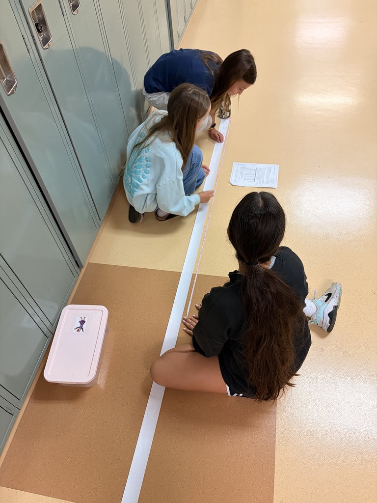Three students sitting in a school hallway analyzing a long paper tape measure during a science lab.