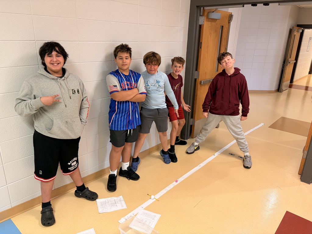 Five students posing together in a school hallway beside a paper measurement line for a science project.