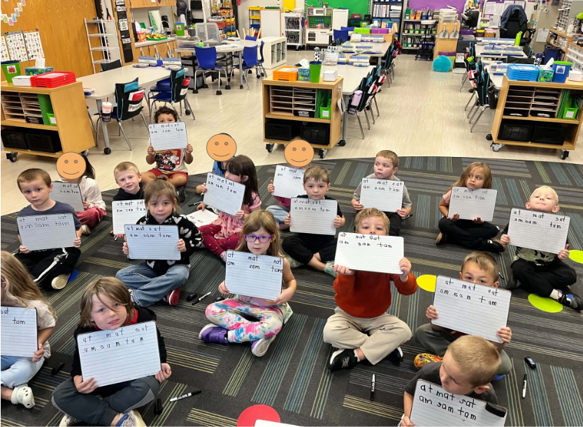 students sitting on carpet in kindergarten room showing the white boards that they used to practice writing words in phonics lesson