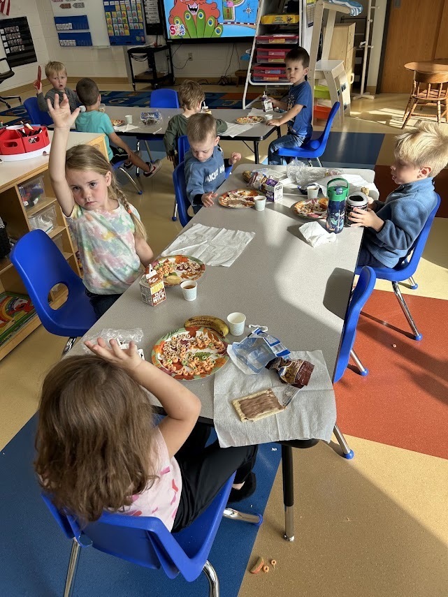 Young students sitting at a classroom table raising their hands during a tasting lesson