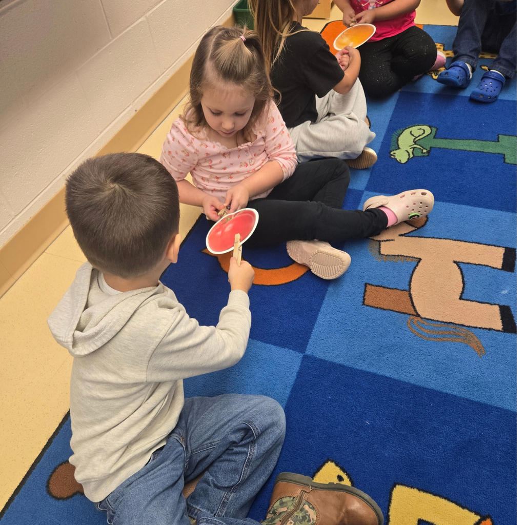 Elementary students sitting on a rug in a classroom, attaching clothespins to paper fruit cutouts during an activity.
