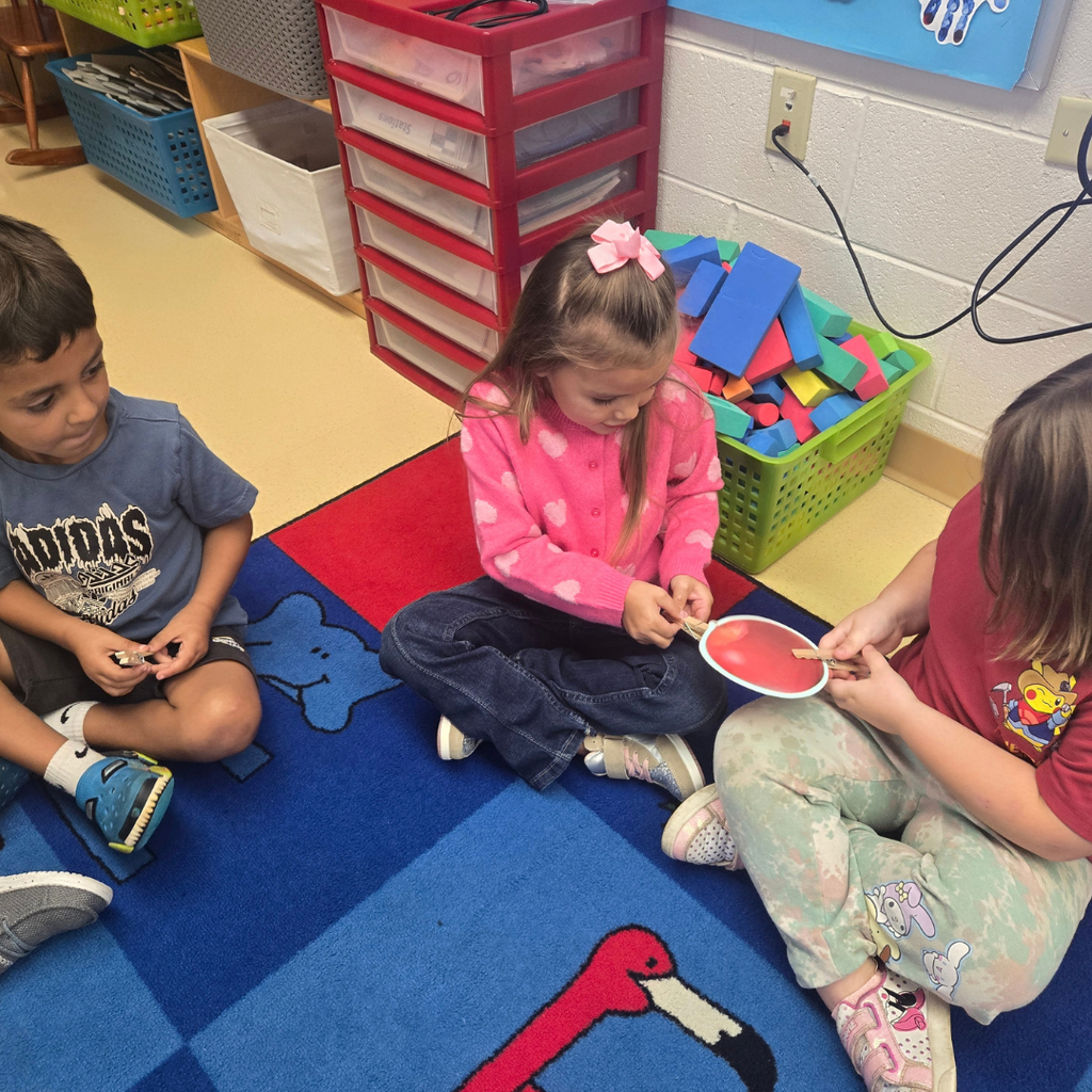 Children sitting on a classroom carpet attaching clothespins to a paper cutout during a group fine-motor skill lesson.