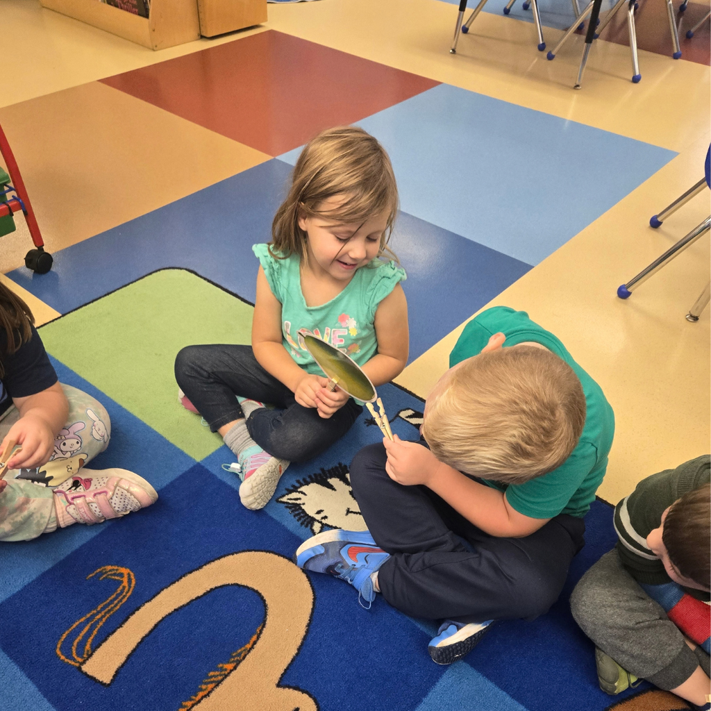 Young students sitting on an alphabet rug working together on a fine-motor skill activity with paper cutouts and clothespins.