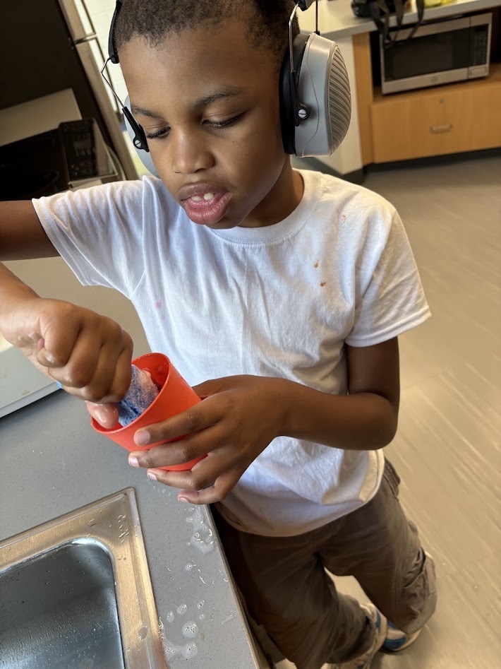 A student wearing noise-canceling headphones cleaning a cup in a sink during a classroom activity